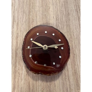 May include: A round clock made from a polished, brown agate slice. The clock face features gold-colored hands and silver dot hour markers. The agate has a natural, layered appearance, creating a unique decorative piece.