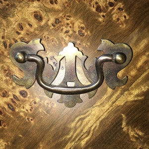 Puede incluir: Un tirador de cajón de latón con un diseño decorativo y ornamentado. El tirador tiene una manija curva y dos piezas laterales decorativas.