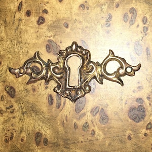 May include: A gold-toned ornate keyhole cover with intricate details and a keyhole in the center. The cover is mounted on a wood surface with a brown and gold mottled pattern.