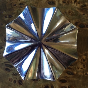 May include: A polished, silver-coloured, octagonal dish with a fluted design. The dish reflects light, creating highlights and shadows. The background is a wood surface with a unique, dark-coloured grain pattern.