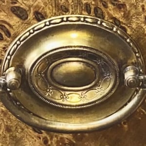 Op de afbeelding: Een gouden metalen kastknop met een sierlijk ontwerp. De knop heeft een verhoogd ovaal midden en een decoratieve rand.