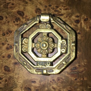 May include: A gold-toned metal cabinet knob with an intricate design. The knob has a square shape with rounded corners and a decorative centre piece.