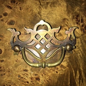 May include: A brass cabinet pull with a decorative, ornate design. The pull has a diamond-shaped grid pattern in the center and a curved, looped handle.