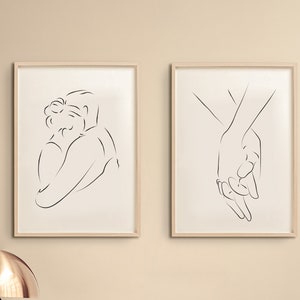 May include: Two framed black and white line drawings of a couple. One drawing shows a couple embracing, the other shows two hands holding each other. The drawings are hanging on a beige wall above a wooden dresser with a lamp and a decorative coral piece on top.