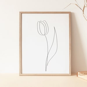 Puede incluir: Una impresi&oacute;n enmarcada con un dibujo lineal minimalista de un tulip&aacute;n. La obra de arte est&aacute; en un marco de madera clara, exhibida sobre una superficie beige. Un jarr&oacute;n de madera con hierba seca y un libro tambi&eacute;n est&aacute;n en la superficie.
