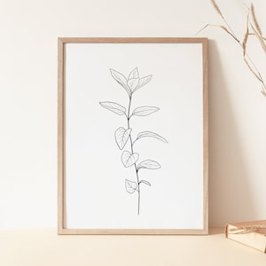 May include: A framed black and white line drawing of a plant with leaves. The frame is light brown wood and the print is on white paper. The print is sitting on a light brown surface.