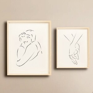 May include: Two minimalist line art prints in wooden frames, one depicting a couple embracing and the other showing two hands holding each other. The prints are displayed on a beige wall above a wooden cabinet.