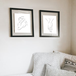May include: Two black framed prints of line drawings depicting a couple embracing and two hands holding each other. The prints are hanging on a white wall above a white bed with gray and white patterned pillows.
