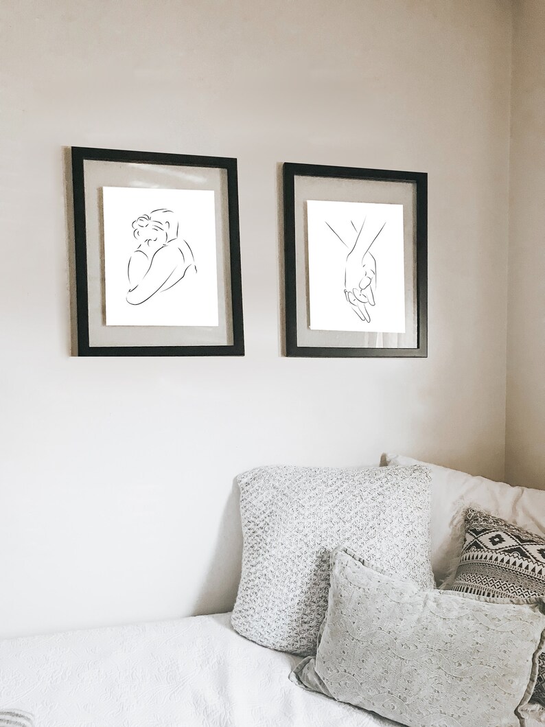 May include: Two black framed prints of line drawings depicting a couple embracing and two hands holding each other. The prints are hanging on a white wall above a white bed with gray and white patterned pillows.