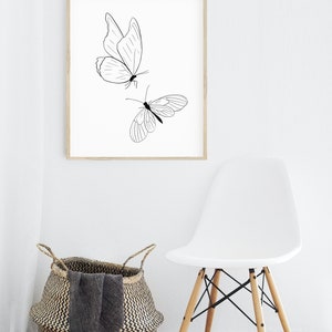 May include: A black and white line drawing of two butterflies on a white background, framed in a light wood frame. The frame is hanging on a white wall above a white chair with wooden legs.