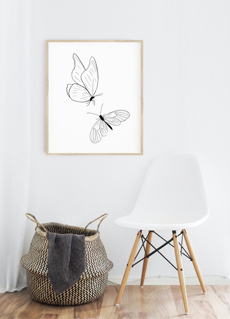 May include: A black and white line drawing of two butterflies on a white background, framed in a light wood frame. The frame is hanging on a white wall above a white chair with wooden legs.