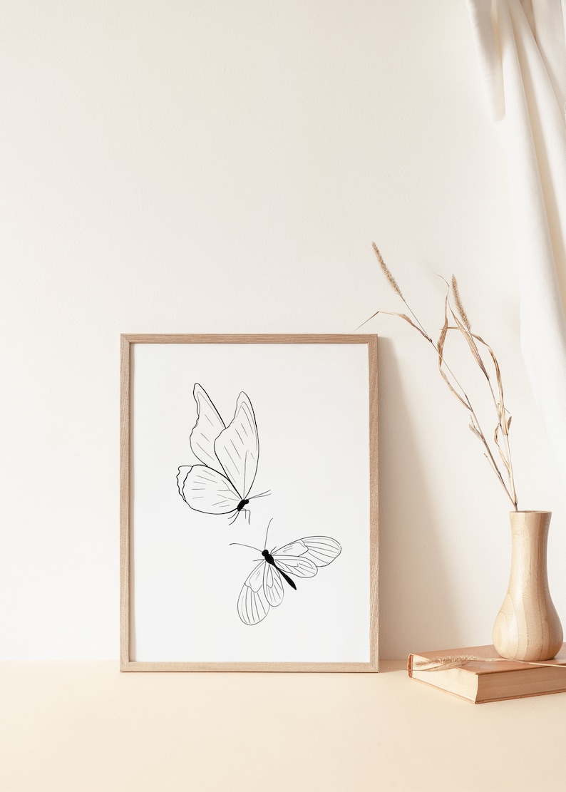 May include: A framed print with a minimalist line drawing of two butterflies in black on a white background. The frame is light wood and the print is sitting on a light beige surface.