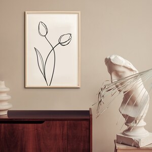 Puede incluir: Un dibujo en blanco y negro de dos tulipes sobre un fondo blanco, enmarcado en un marco de madera clara. El dibujo est&aacute; colgado en una pared beige. Un jarr&oacute;n de cer&aacute;mica blanco se encuentra sobre una mesa de madera oscura en primer plano. Un busto de mujer de yeso blanco se encuentra sobre un pedestal de madera a la derecha del marco.