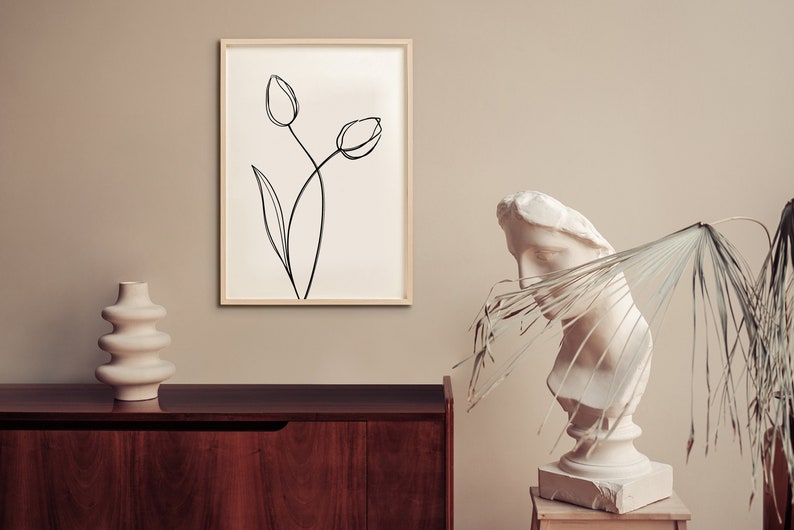 Puede incluir: Un dibujo en blanco y negro de dos tulipes sobre un fondo blanco, enmarcado en un marco de madera clara. El dibujo est&aacute; colgado en una pared beige. Un jarr&oacute;n de cer&aacute;mica blanco se encuentra sobre una mesa de madera oscura en primer plano. Un busto de mujer de yeso blanco se encuentra sobre un pedestal de madera a la derecha del marco.