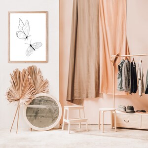 May include: A framed black and white line drawing of two butterflies on a white background. The frame is hanging on a white wall. The wall is next to a room with a wooden clothes rack, two wooden stools, and a round mirror.