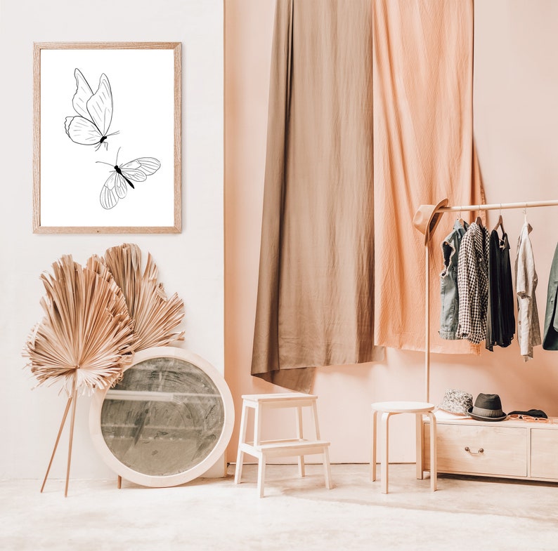 May include: A framed black and white line drawing of two butterflies on a white background. The frame is hanging on a white wall. The wall is next to a room with a wooden clothes rack, two wooden stools, and a round mirror.