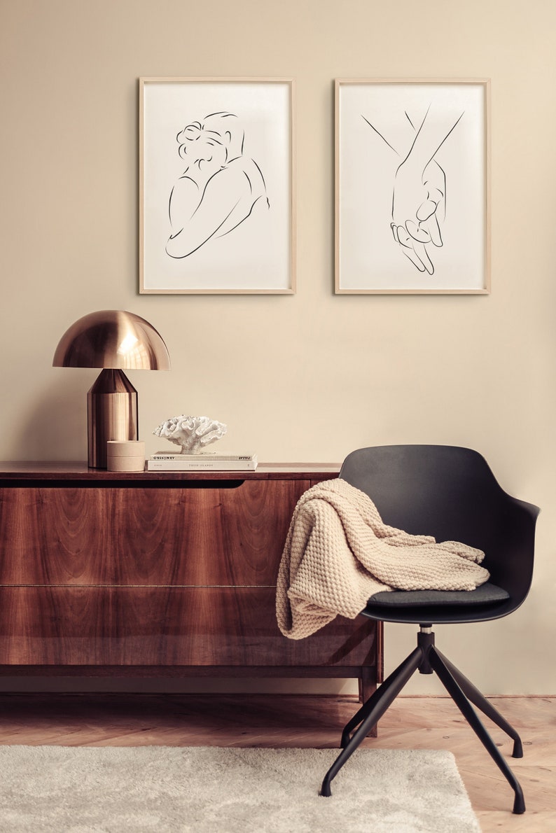 May include: Two framed black and white line drawings of a couple. One drawing shows a couple embracing, the other shows two hands holding each other. The drawings are hanging on a beige wall above a wooden dresser with a lamp and a decorative coral piece on top.