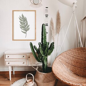 May include: A white wooden dresser with three drawers, a wicker chair, a potted cactus, a round white rug with pom-poms, a lantern, and a framed print of a green leaf.