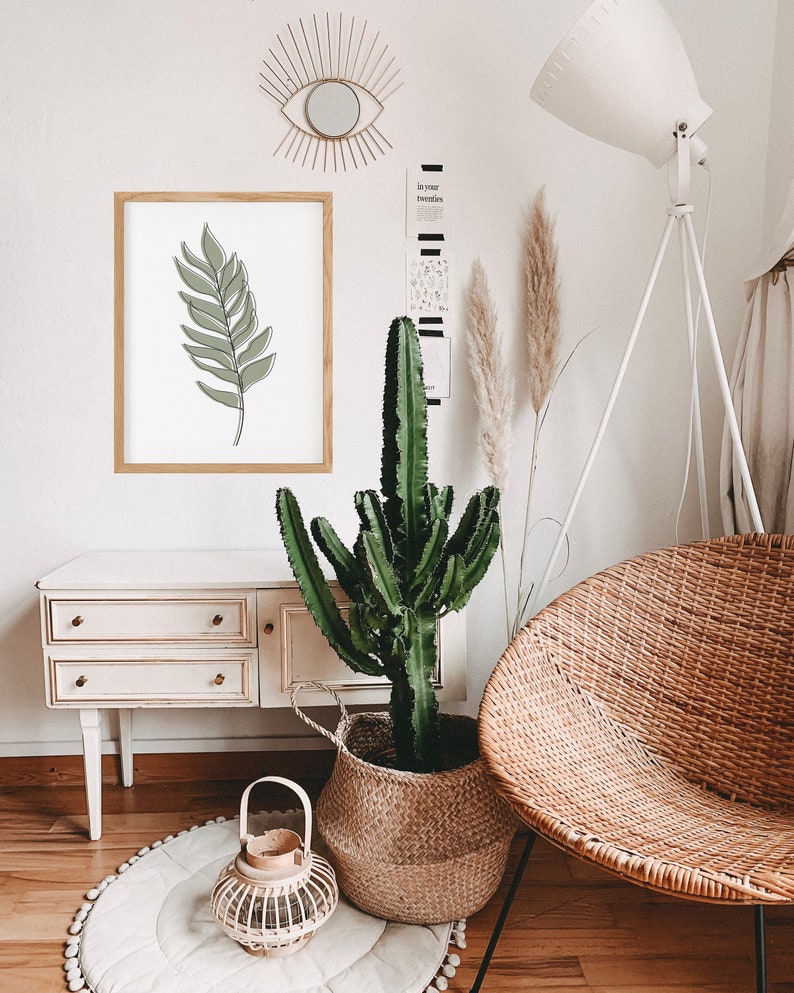 May include: A white wooden dresser with three drawers, a wicker chair, a potted cactus, a round white rug with pom-poms, a lantern, and a framed print of a green leaf.
