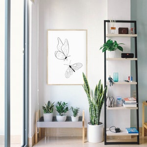 May include: A black and white line drawing of two butterflies on a white background in a wooden frame. The frame is hanging on a white wall. There are plants in pots on a shelf below the frame.