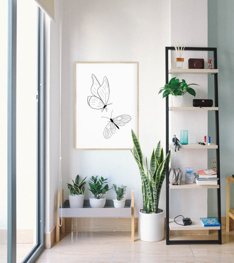 May include: A black and white line drawing of two butterflies on a white background in a wooden frame. The frame is hanging on a white wall. There are plants in pots on a shelf below the frame.