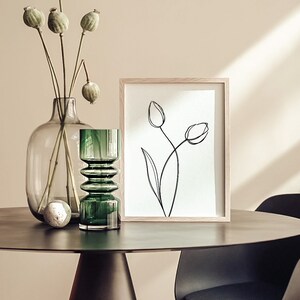 Puede incluir: Un dibujo en blanco y negro de dos tulipes en un marco de madera, sentado en una mesa con un jarr&oacute;n de vidrio verde y una botella de vidrio verde con una esfera blanca dentro.