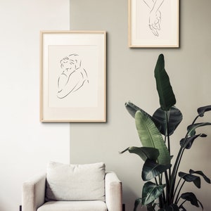 May include: Two framed line drawings of figures in a minimalist style. The drawing on the left shows two people embracing. The drawing on the right shows two hands holding each other. The drawings are hung on a light beige wall above a white armchair. A large green plant in a gold pot is in the corner of the room.