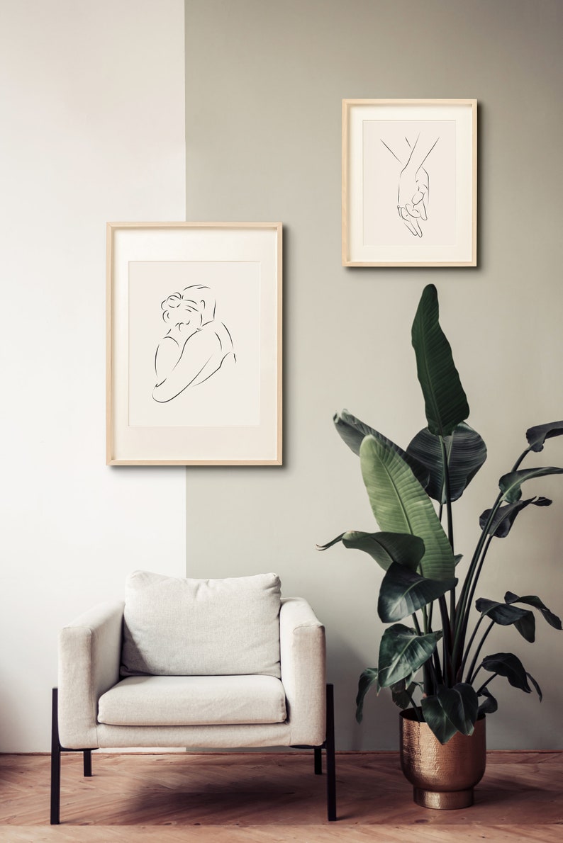 May include: Two framed line drawings of figures in a minimalist style. The drawing on the left shows two people embracing. The drawing on the right shows two hands holding each other. The drawings are hung on a light beige wall above a white armchair. A large green plant in a gold pot is in the corner of the room.