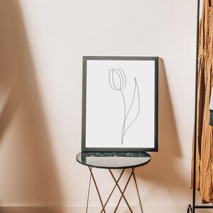 Puede incluir: Un dibujo lineal en blanco y negro enmarcado de un tulip&aacute;n. La obra de arte se exhibe sobre una peque&ntilde;a mesa redonda con tapa verde y patas doradas. En el fondo se ve un perchero con varias prendas.
