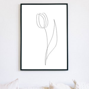 Puede incluir: L&aacute;mina art&iacute;stica enmarcada en negro con un dibujo lineal minimalista de una flor de tulip&aacute;n. La obra de arte es un dise&ntilde;o sencillo y elegante con una sola l&iacute;nea continua que forma la flor y el tallo sobre un fondo blanco. La impresi&oacute;n se muestra sobre un sof&aacute; blanco.