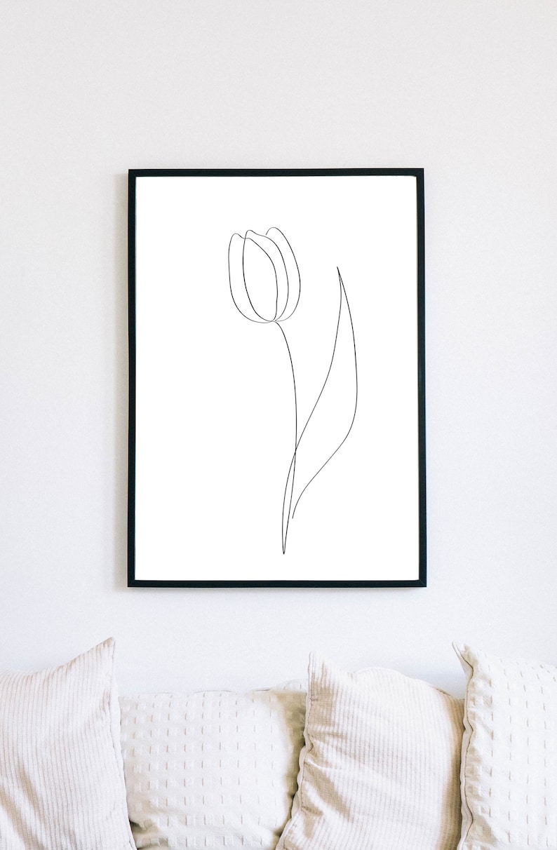 Puede incluir: L&aacute;mina art&iacute;stica enmarcada en negro con un dibujo lineal minimalista de una flor de tulip&aacute;n. La obra de arte es un dise&ntilde;o sencillo y elegante con una sola l&iacute;nea continua que forma la flor y el tallo sobre un fondo blanco. La impresi&oacute;n se muestra sobre un sof&aacute; blanco.
