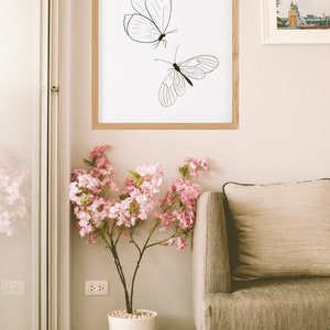 May include: A framed print of two black line drawings of butterflies on a white background. The print is hanging on a light beige wall above a brown sofa and a potted pink flowering plant.