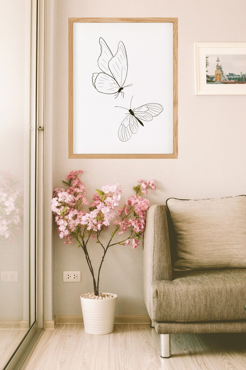 May include: A framed print of two black line drawings of butterflies on a white background. The print is hanging on a light beige wall above a brown sofa and a potted pink flowering plant.