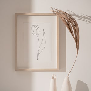 Puede incluir: Una impresi&oacute;n art&iacute;stica minimalista enmarcada con un dibujo lineal negro de un tulip&aacute;n. La obra de arte est&aacute; en un marco de madera clara, exhibida sobre una superficie blanca con jarrones decorativos y una hoja de palma seca. La est&eacute;tica general es limpia y moderna.