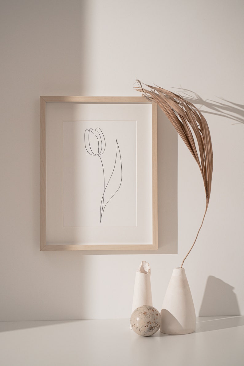 Puede incluir: Una impresi&oacute;n art&iacute;stica minimalista enmarcada con un dibujo lineal negro de un tulip&aacute;n. La obra de arte est&aacute; en un marco de madera clara, exhibida sobre una superficie blanca con jarrones decorativos y una hoja de palma seca. La est&eacute;tica general es limpia y moderna.