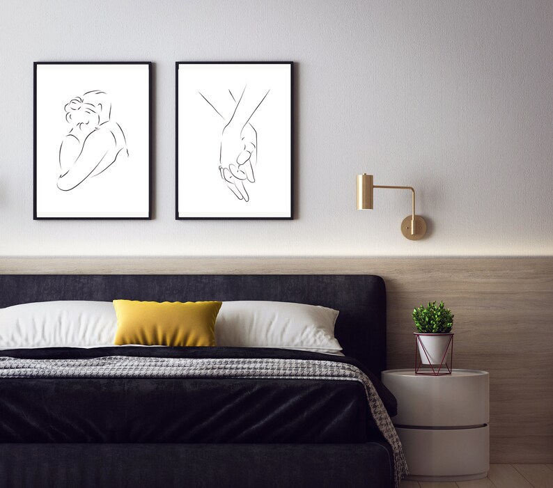 May include: Two black framed line art prints depicting a couple embracing and two hands holding each other. The prints are hanging on a white wall above a black headboard bed with a yellow throw pillow.