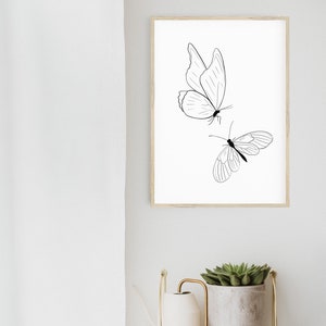 May include: A black and white line drawing of two butterflies on a white background. The butterflies are in a simple, minimalist style. The drawing is framed in a light wood frame.