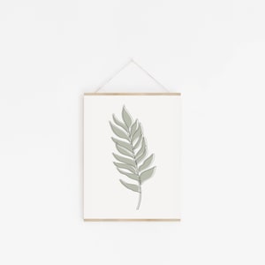 May include: A framed print of a single green leaf with a simple line drawing style. The leaf is hanging on a white wall.