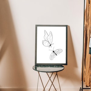 May include: A black framed print with a white background featuring a line drawing of two butterflies. The print is displayed on a black side table with a gold metal base.