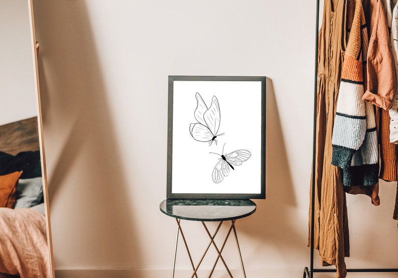 May include: A black framed print with a white background featuring a line drawing of two butterflies. The print is displayed on a black side table with a gold metal base.