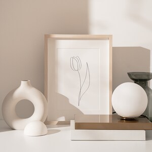 Puede incluir: Naturaleza muerta minimalista con un dibujo lineal de tulip&aacute;n enmarcado, una l&aacute;mpara de globo blanca y un jarr&oacute;n decorativo. La obra de arte est&aacute; en un marco de madera clara. Otros objetos incluyen un jarr&oacute;n de cer&aacute;mica blanca con una abertura circular.