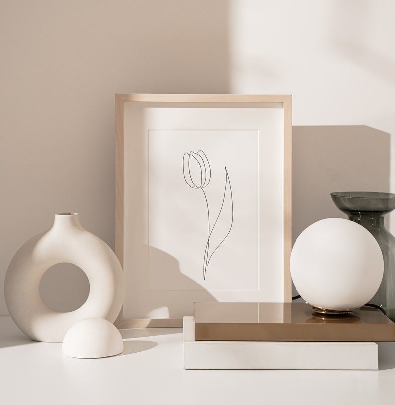 Puede incluir: Naturaleza muerta minimalista con un dibujo lineal de tulip&aacute;n enmarcado, una l&aacute;mpara de globo blanca y un jarr&oacute;n decorativo. La obra de arte est&aacute; en un marco de madera clara. Otros objetos incluyen un jarr&oacute;n de cer&aacute;mica blanca con una abertura circular.