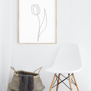 Puede incluir: Un cuadro con una impresi&oacute;n art&iacute;stica minimalista de un dibujo lineal negro de un tulip&aacute;n. La obra de arte est&aacute; colgada en una pared blanca sobre una cesta tejida y una moderna silla blanca con patas de madera. La habitaci&oacute;n tiene un ambiente luminoso y aireado.