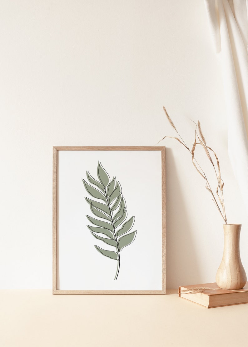 May include: A framed print of a single green leaf drawn in a minimalist line art style. The print is in a light wood frame and is sitting on a table with a vase of dried grasses.