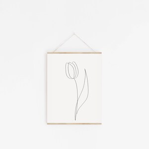Puede incluir: Una impresi&oacute;n art&iacute;stica minimalista que presenta un dibujo de una sola l&iacute;nea de un tulip&aacute;n. El contorno negro de la flor y el tallo se establece sobre un fondo crema. La obra de arte est&aacute; enmarcada con un colgador de madera natural y suspendida por un cord&oacute;n de color neutro.