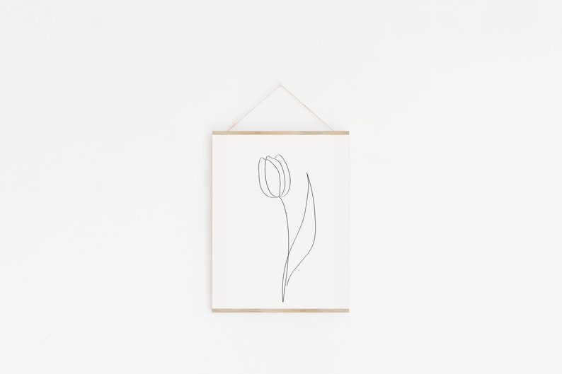 Puede incluir: Una impresi&oacute;n art&iacute;stica minimalista que presenta un dibujo de una sola l&iacute;nea de un tulip&aacute;n. El contorno negro de la flor y el tallo se establece sobre un fondo crema. La obra de arte est&aacute; enmarcada con un colgador de madera natural y suspendida por un cord&oacute;n de color neutro.