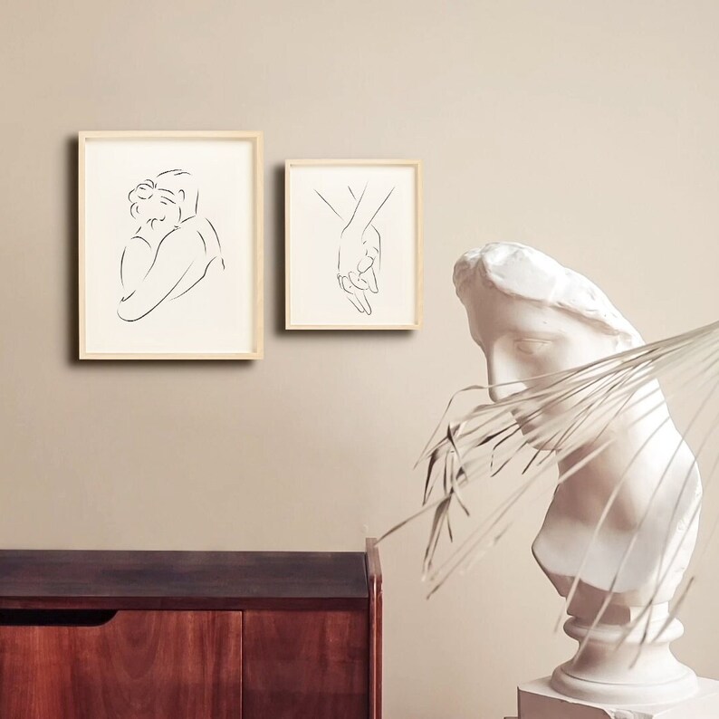 May include: Two minimalist line art prints in wooden frames, one depicting a couple embracing and the other showing two hands holding each other. The prints are displayed on a beige wall above a wooden cabinet.