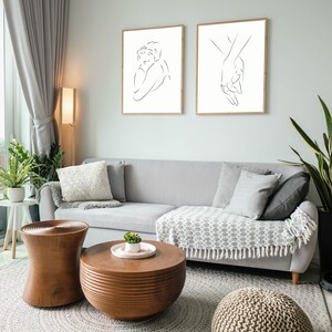 May include: Two black and white line art prints of a couple embracing and two hands holding each other. The prints are framed in natural wood and hung on a light gray wall above a gray couch with pillows and a throw blanket. The couch is in a living room with a wooden coffee table and a round ottoman.