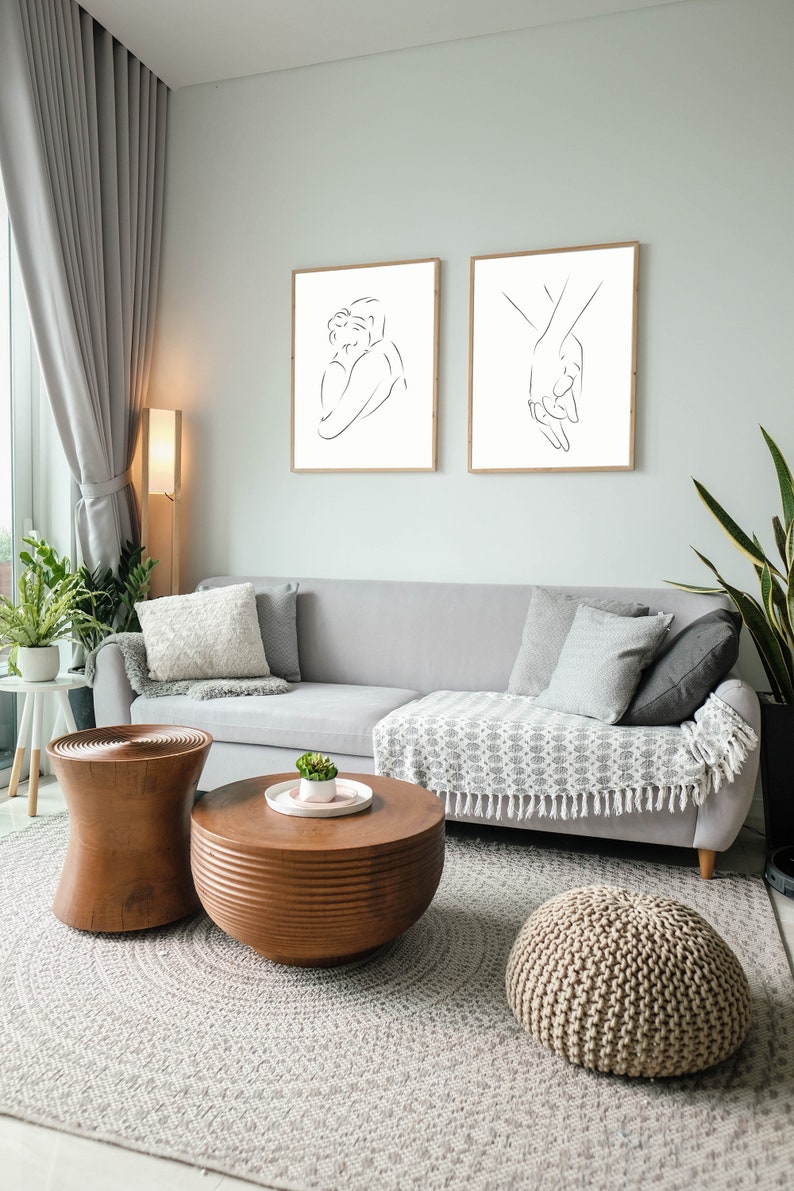 May include: Two black and white line art prints of a couple embracing and two hands holding each other. The prints are framed in natural wood and hung on a light gray wall above a gray couch with pillows and a throw blanket. The couch is in a living room with a wooden coffee table and a round ottoman.