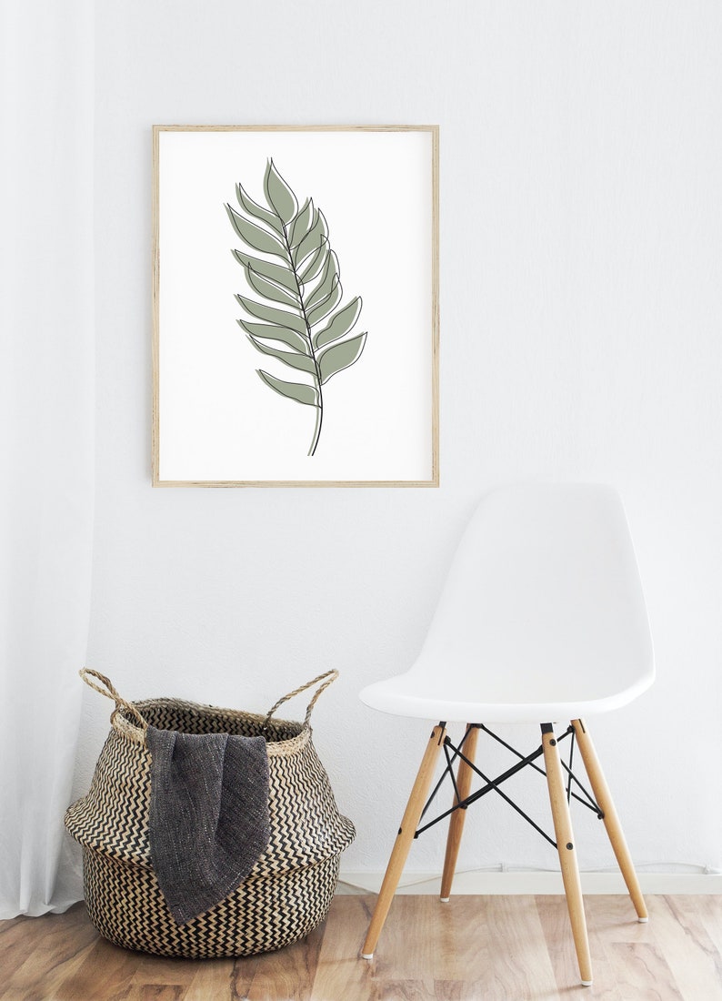 May include: A framed print of a single green leaf drawn in a minimalist style. The print is hanging on a white wall above a white chair and a woven basket.